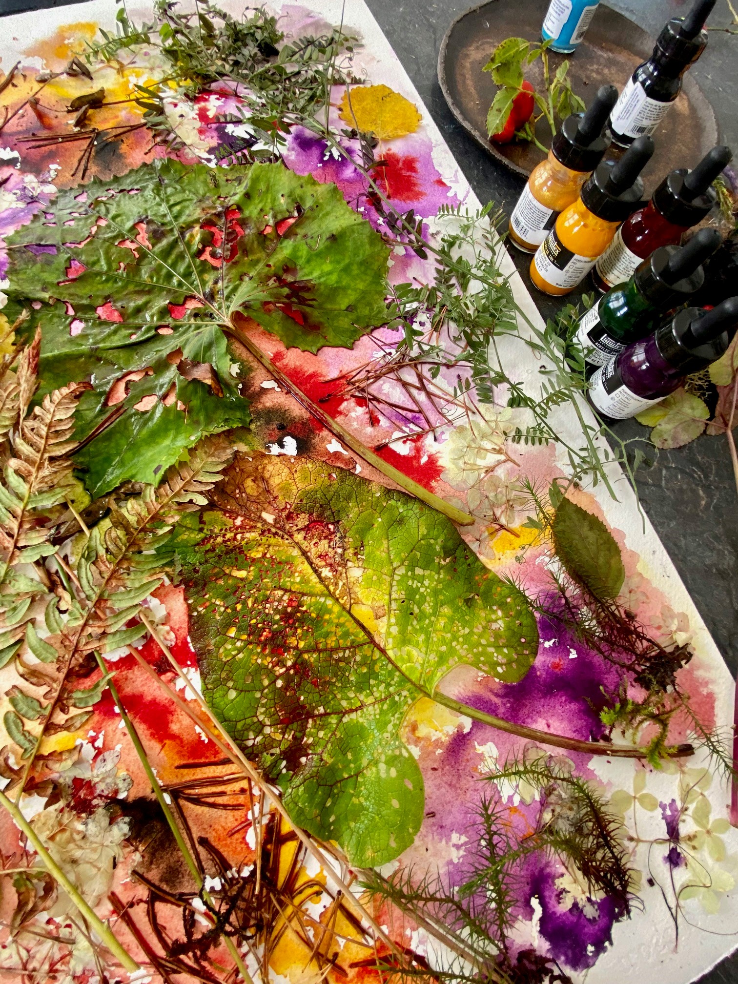 Kunstiteos, mis koosneb erinevatest taimedest, õitest ja värvilaikudest. Pildi ülemises osas on näha  värvipudelid.